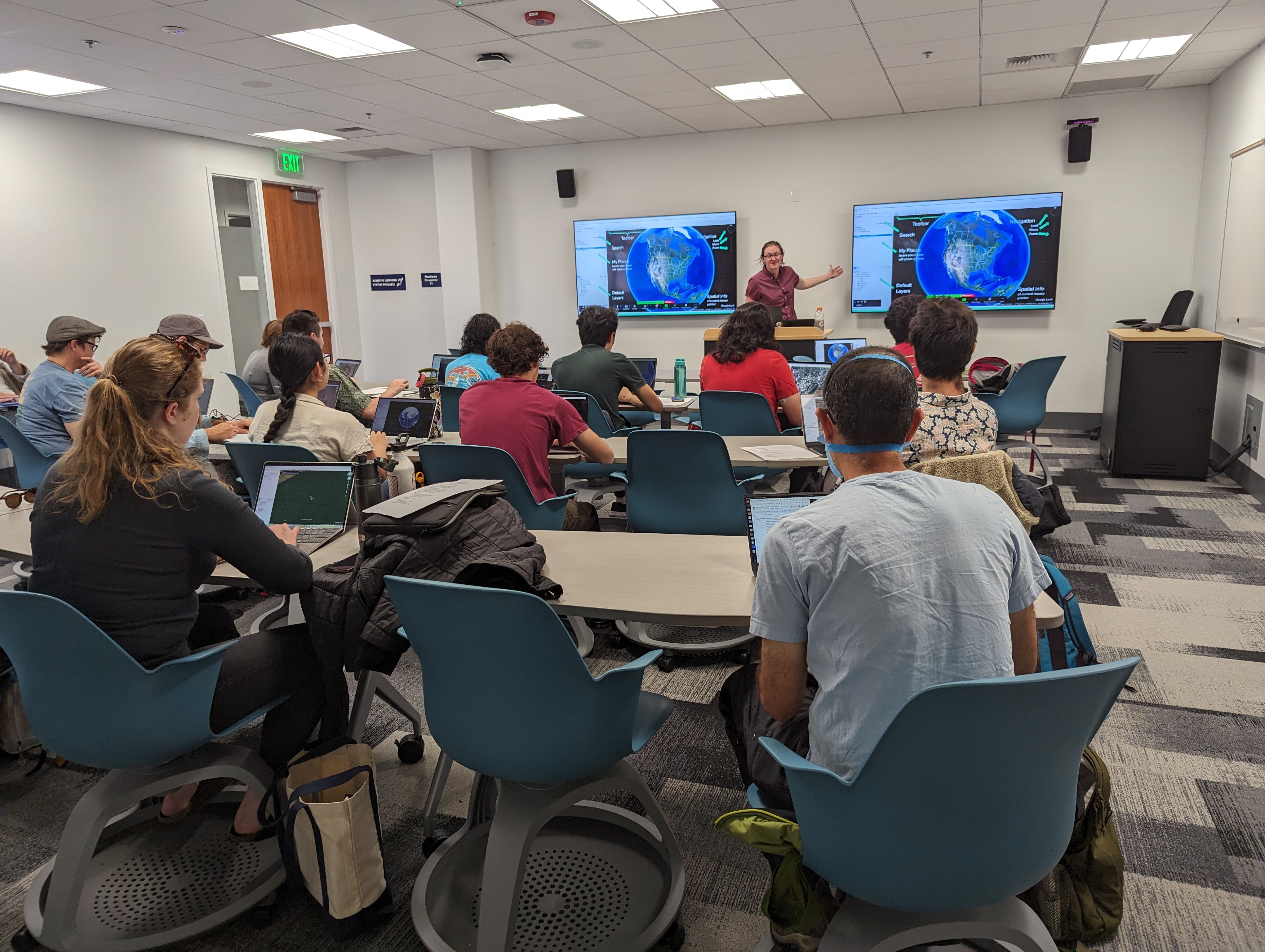 Taken from the back of the room at Physical and Data Sciences Building Seminar Room where students sit in blue chairs at rows of long tables facing a podium and digital screens where an instructor points at a globe on one of the two screens. 